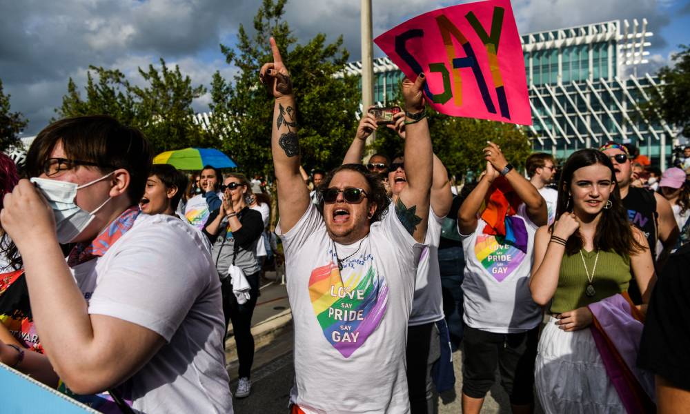 A person wearing a white shirt with a rainbow heart on it shouts as they hold up a pink sign that reads "GAY" during a protest against Florida's 'Don't Say Gay' legislation