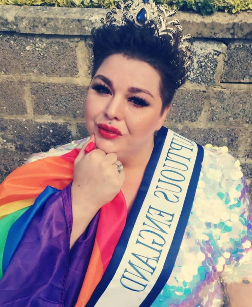 In this photo, Laura Davis has a rainbow LGBTQ+ Pride flag draped over one arm as she wears a sequinned dress. She is also wearing a beauty pageant sash and crown