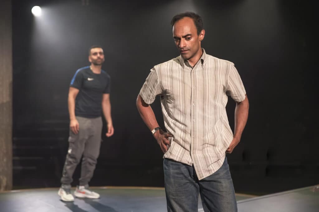 Billy, in the background, wearing sportswear, Zafar in the foreground in a shirt and jeans looking troubled